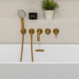 Howesto Sp z o.o. - Minimalistyczna łazienka: biała wanna z armaturą w kolorze szczotkowanego złota, półka z rośliną i świecą. Eleganckie wykończenie marmurowej ściany.