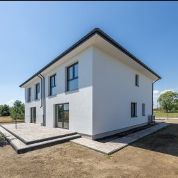 Howesto Sp z o.o. - Nowoczesny, dwukondygnacyjny dom z białym tynkiem i ciemną stolarką okienną, otoczony trawnikiem i drzewami. Widoczny taras z kostki brukowej.