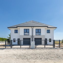 Howesto Sp z o.o. - Nowoczesny, dwukondygnacyjny bliźniak z szarą dachówką i czarnymi oknami, ogrodzony metalową siatką na tle błękitnego nieba. Widoczne przyłącze elektryczne.