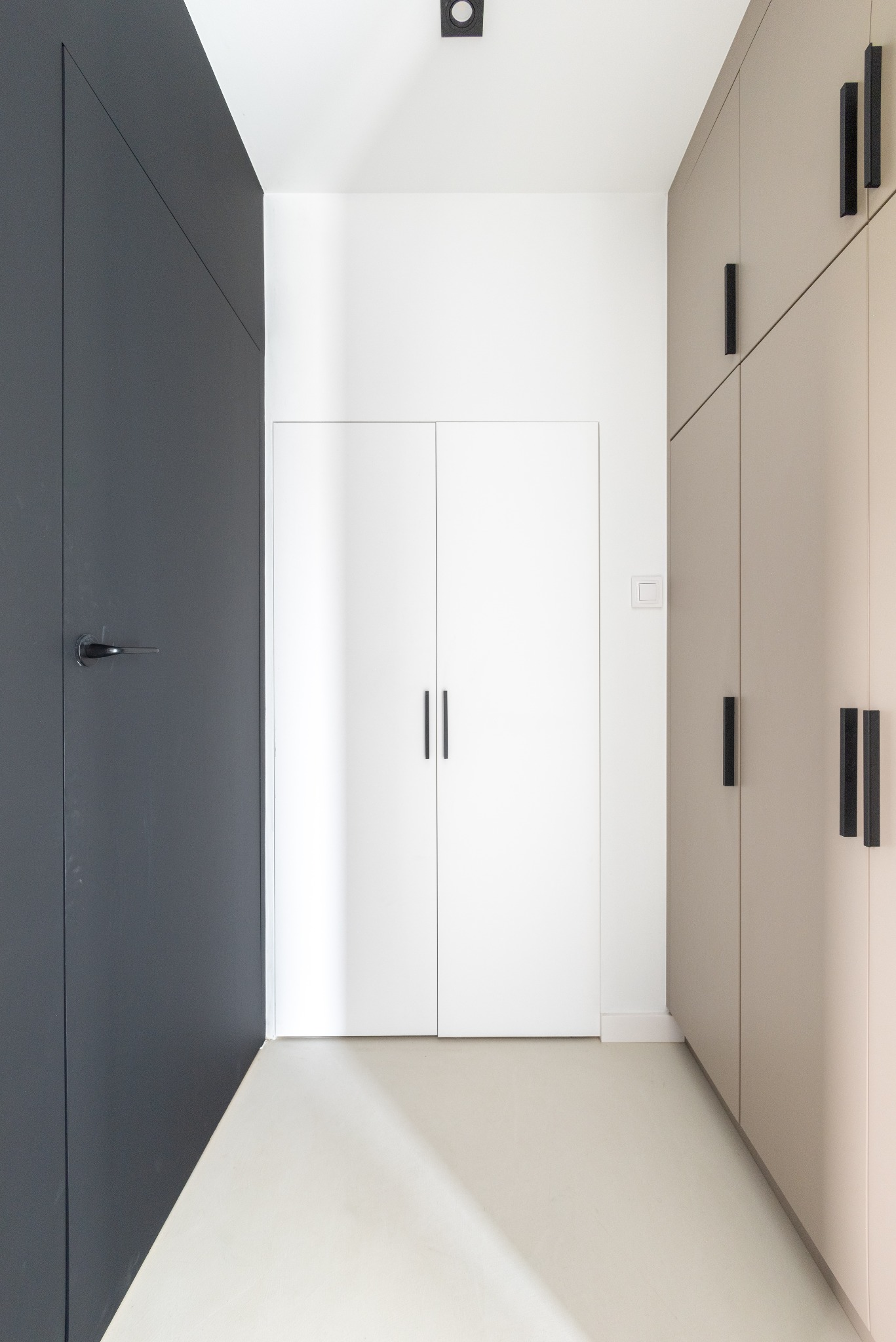 Minimalistyczne wnętrze: antracytowe drzwi z klamką, białe drzwi przesuwne z czarnymi uchwytami i beżowa zabudowa szafy z podłużnymi uchwytami.