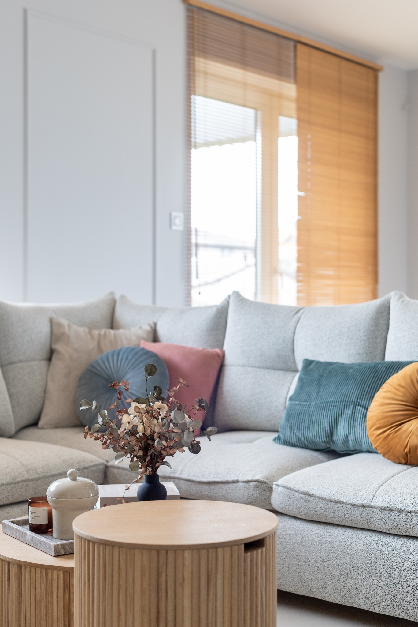 Jasny salon z narożnikiem, dekoracyjnymi poduszkami i okrągłymi stolikami kawowymi. Na stoliku wazon z suszem i świeca. W tle okno z drewnianymi żaluzjami.