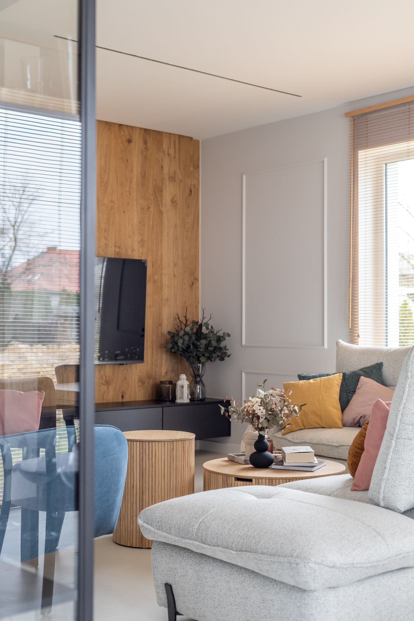 Stylowy salon w Warszawie z drewnianą ścianą TV, szarą sofą z kolorowymi poduszkami i okrągłymi stolikami kawowymi. Widok przez szklane drzwi.