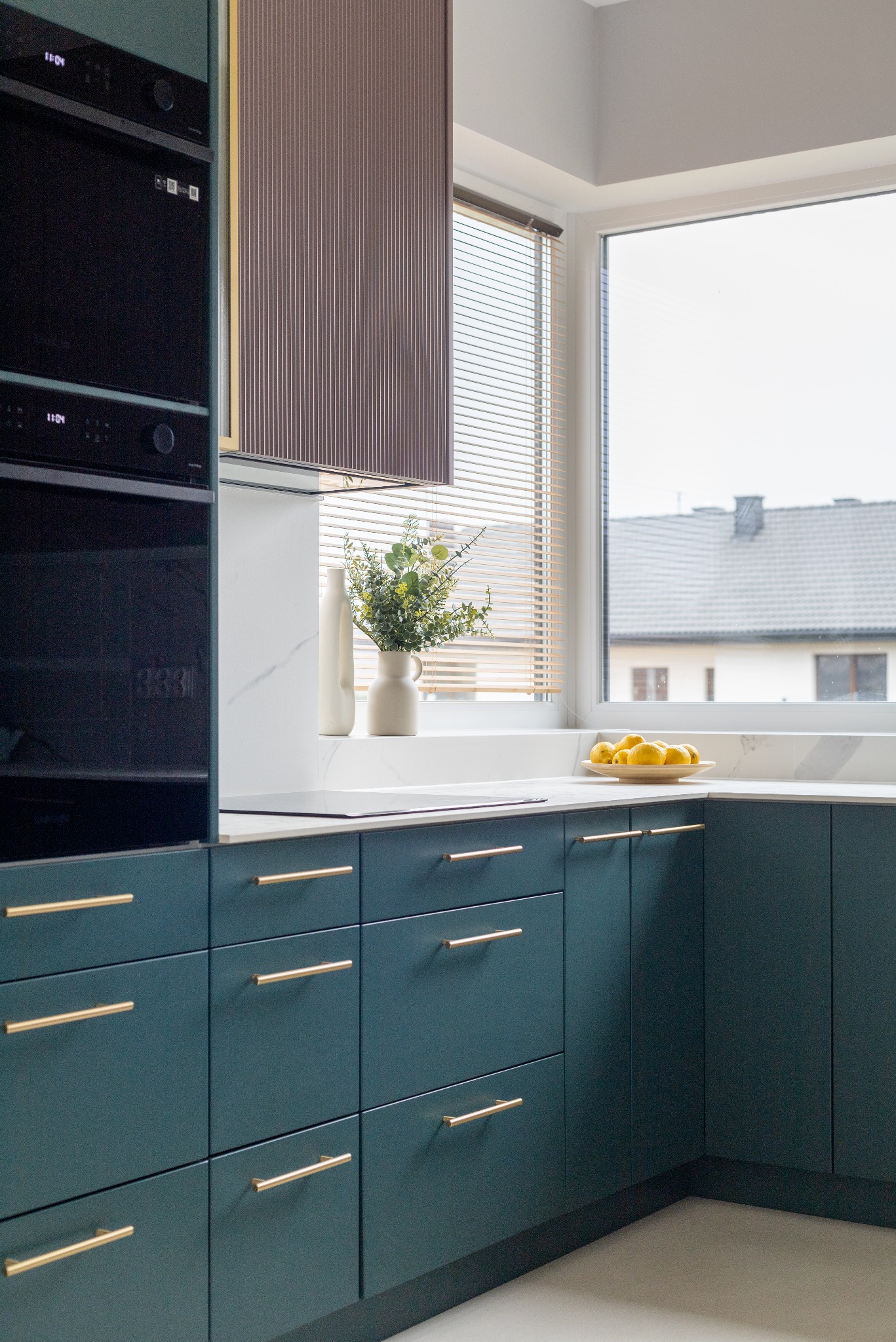 Nowoczesna kuchnia z ciemnozielonymi szafkami, złotymi uchwytami i marmurowym blatem. Na blacie wazon z kwiatami i miska z cytrynami. Widok na okno z żaluzjami.