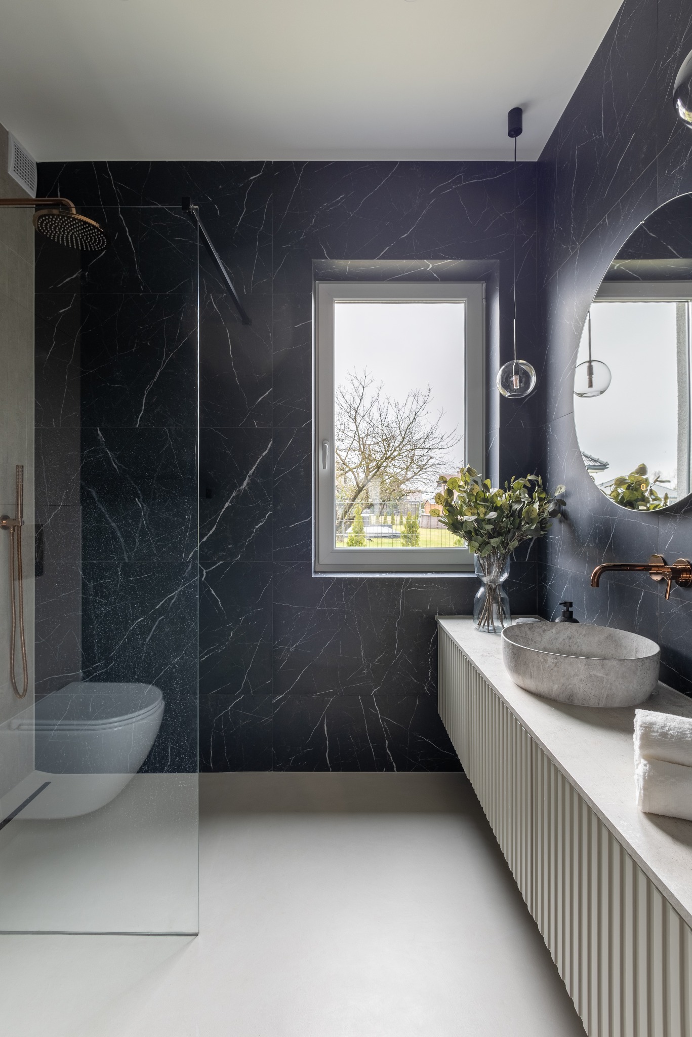 Elegancka łazienka z czarnymi płytkami imitującymi marmur, kabiną prysznicową typu walk-in, owalną umywalką i okrągłym lustrem. Nowoczesny design z akcentami w kolorze miedzi.