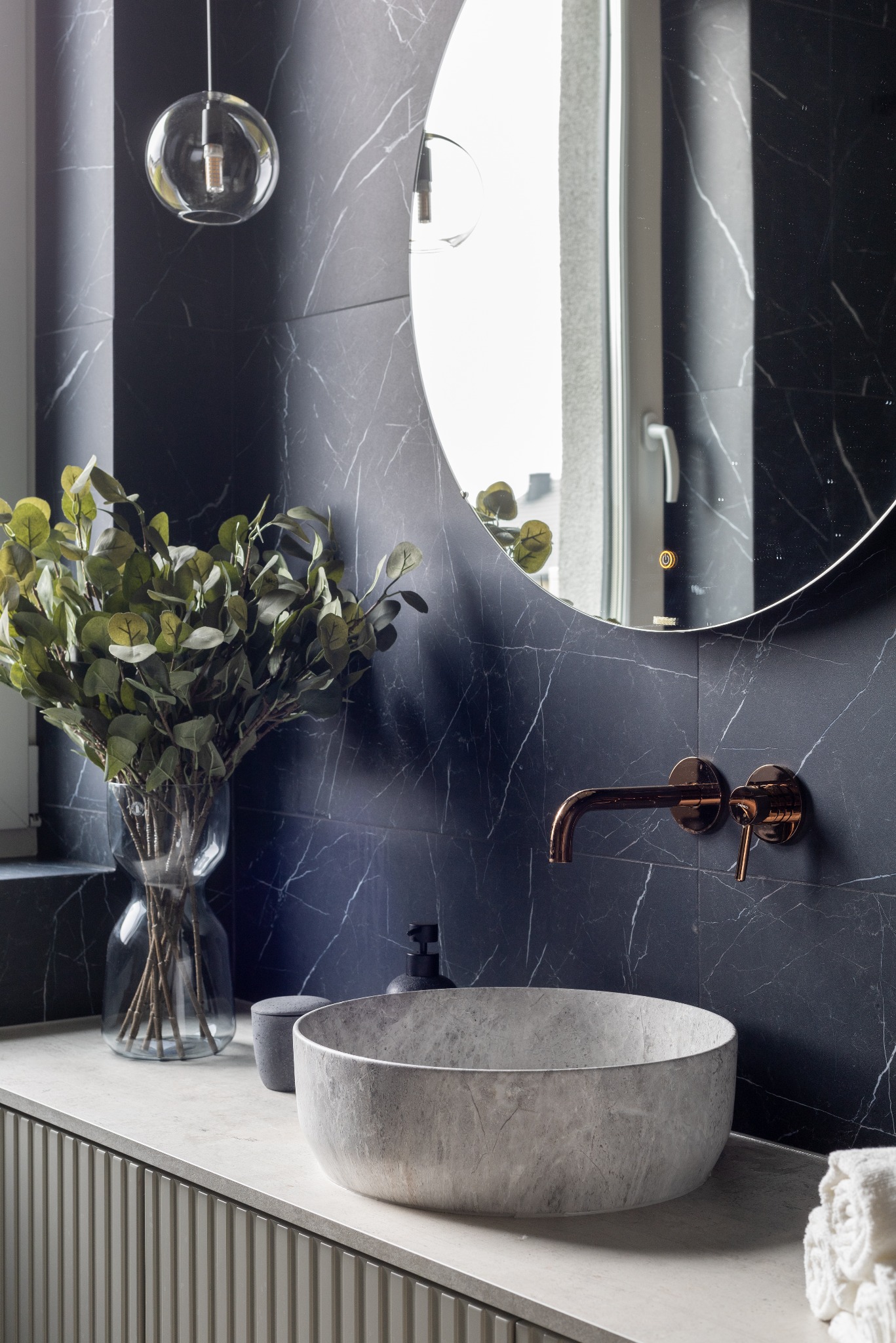 Stylowa łazienka z umywalką z szarego kamienia, miedzianą armaturą i czarnymi płytkami imitującymi marmur. Okrągłe lustro z podświetleniem i szklana lampa.