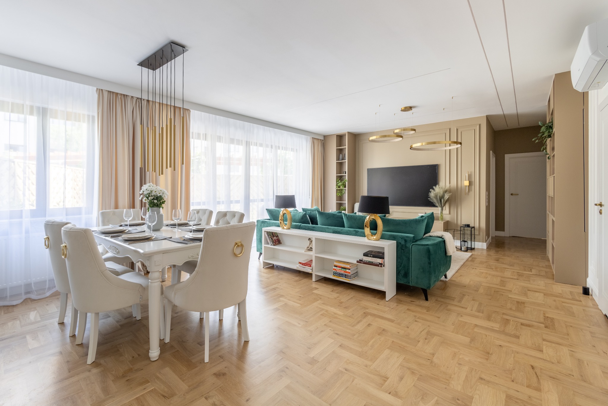 Eleganckie wnętrze: parkiet w jodełkę, stół z nakryciem, krzesła z ozdobnymi uchwytami, zielona sofa i złote lampy. Całość w jasnych, stonowanych barwach.