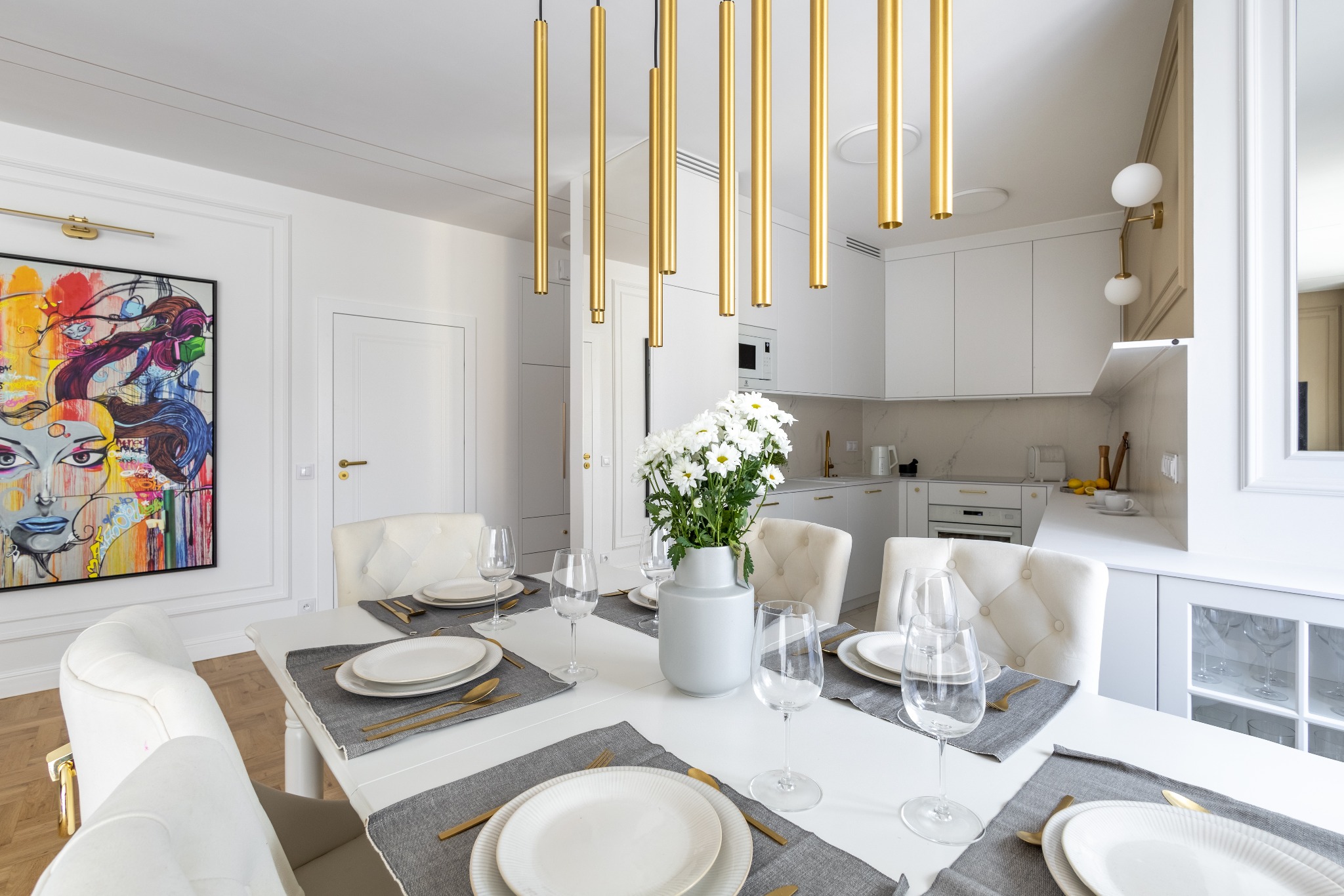 Jasne, nowoczesne wnętrze jadalni z białym stołem, krzesłami i złotymi akcentami oświetleniowymi. W tle widoczna kuchnia z białymi szafkami i marmurowym blatem.