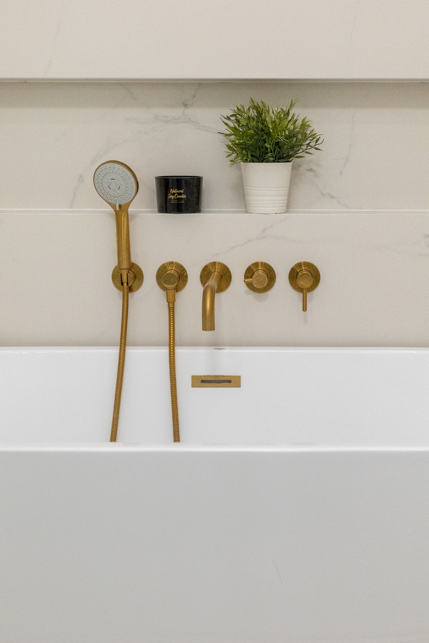 Minimalistyczna łazienka: biała wanna z armaturą w kolorze szczotkowanego złota, półka z rośliną i świecą. Eleganckie wykończenie marmurowej ściany.