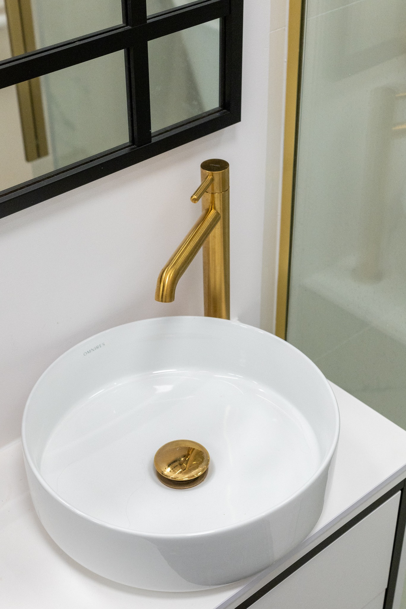 Biała, okrągła umywalka nablatowa z złotą baterią i korkiem, czarna rama lustra w tle. Minimalistyczny design łazienki.