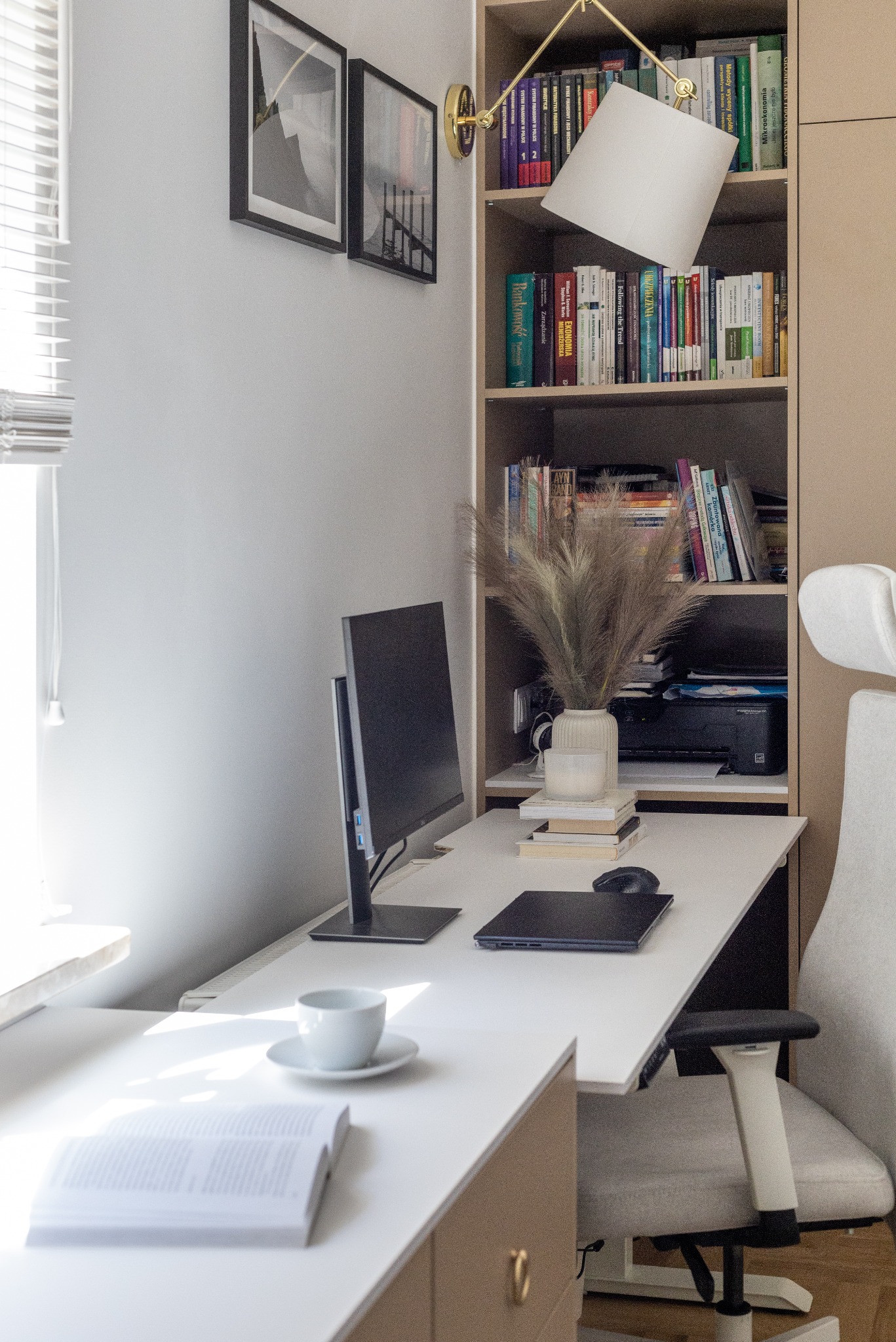 Jasne biuro w domu z białym biurkiem, monitorem, laptopem, książkami i dekoracyjną trawą w wazonie. Nad biurkiem półki z książkami i lampa. Dwa obrazy na ścianie.