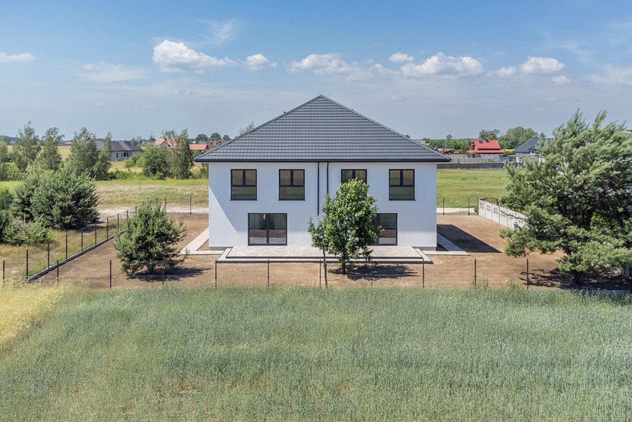 Nowoczesny, dwukondygnacyjny dom z szarą dachówką, białymi ścianami i czarnymi ramami okiennymi, otoczony zielenią i ogrodzeniem z siatki, w słoneczny dzień.