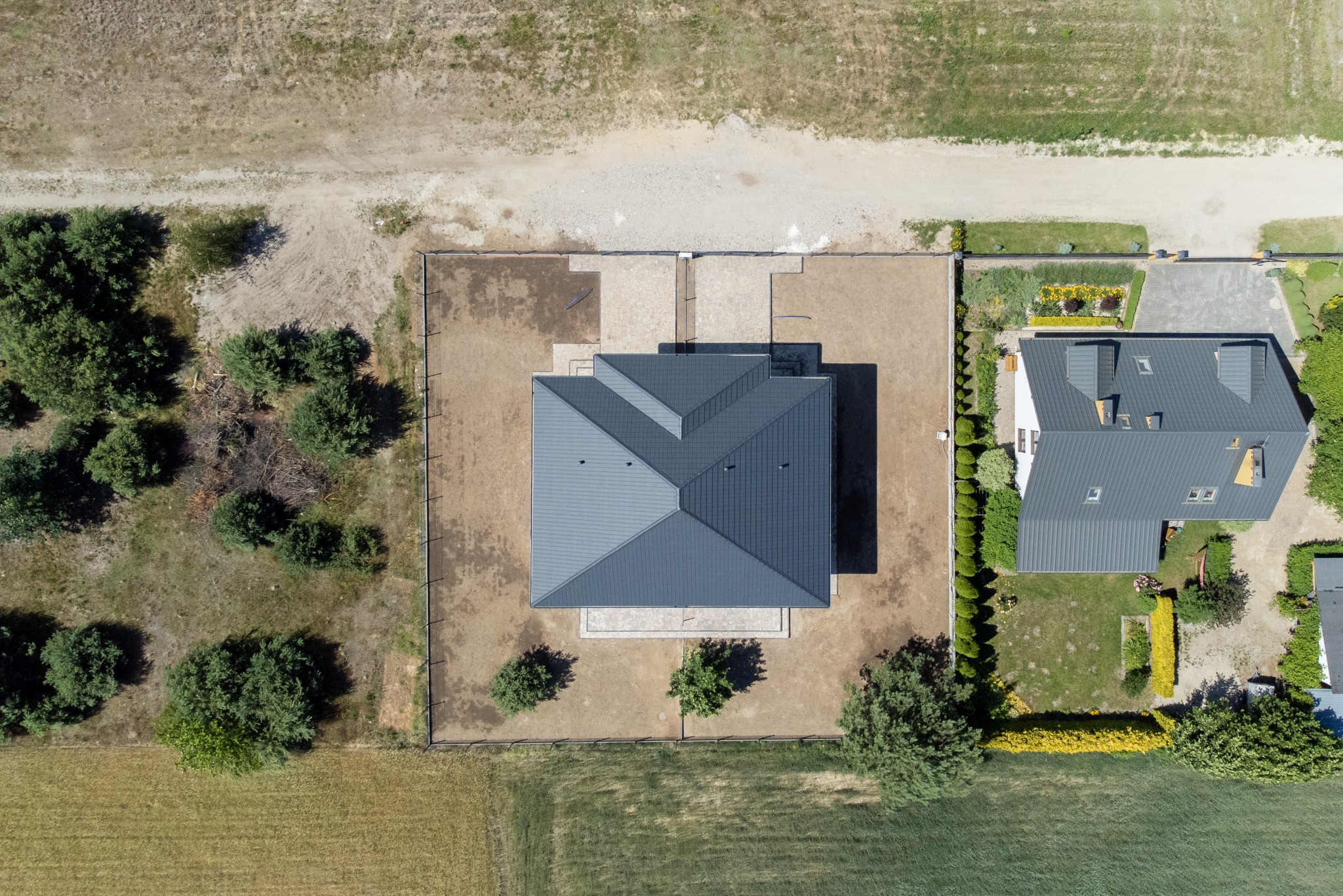 Zdjęcie z lotu ptaka przedstawia dwa domy z szarymi dachami, otoczone zielenią i ogrodzeniami. Widoczne ścieżki i podjazdy z kostki brukowej. Tło stanowią pola i drzewa.