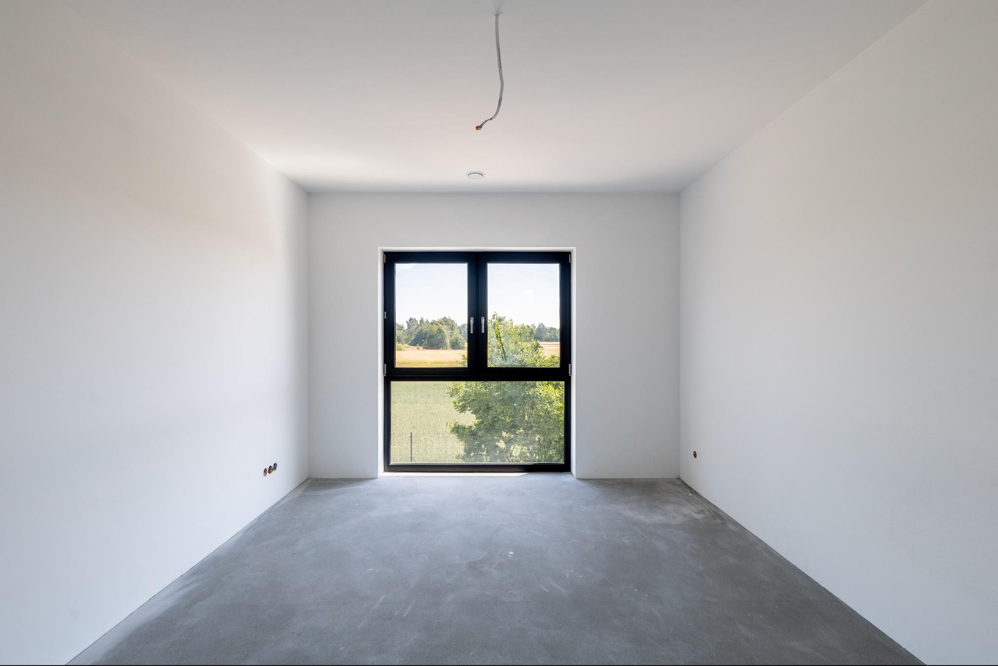 Surowe wnętrze z dużym oknem z czarną ramą, widok na zieleń. Ściany białe, gładkie, podłoga betonowa. Wystające kable elektryczne. Minimalistyczny design.