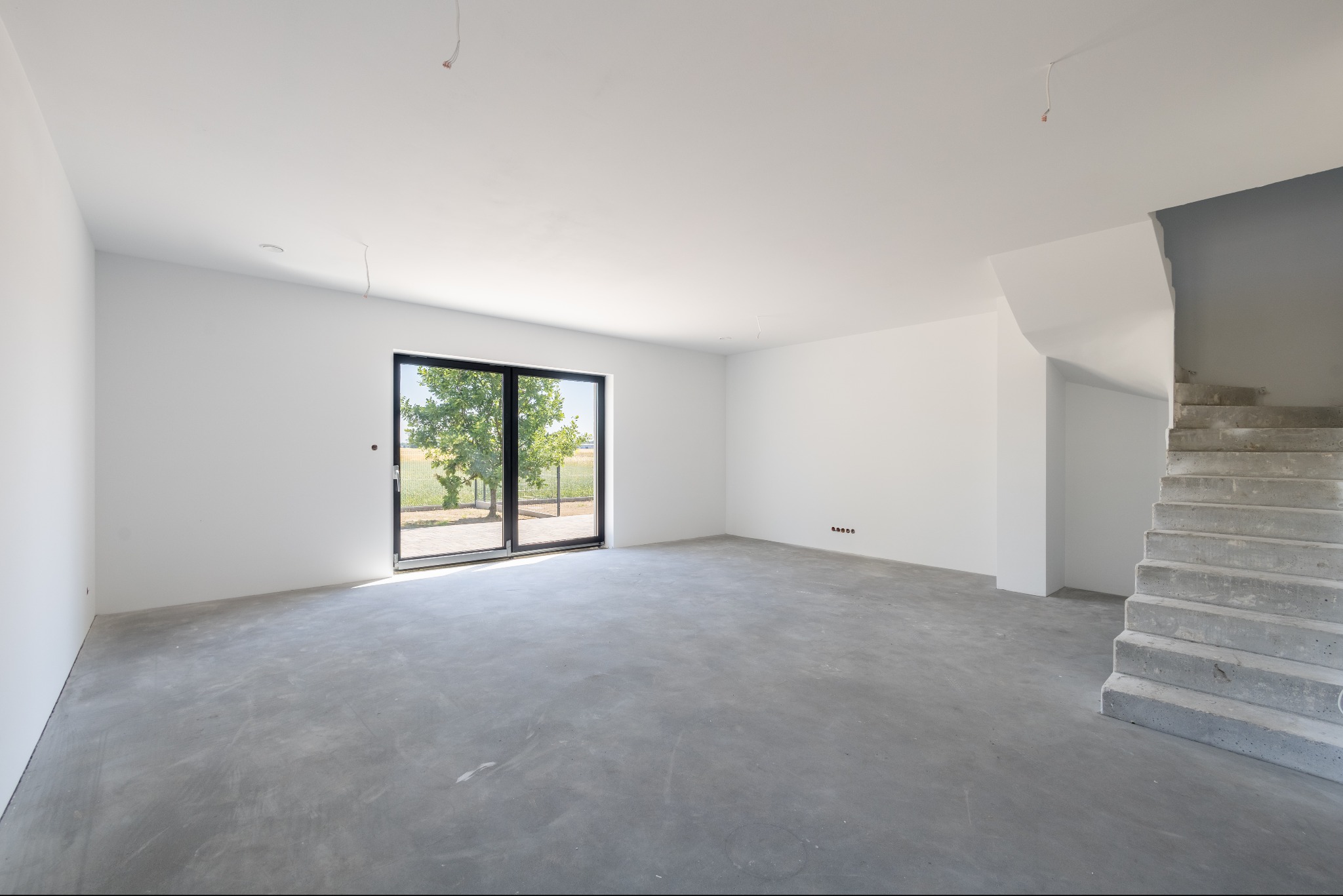 Surowe wnętrze domu w stanie deweloperskim: betonowa podłoga, białe ściany, surowe schody i duże okno z widokiem na zieleń. Widoczne gniazdka elektryczne.