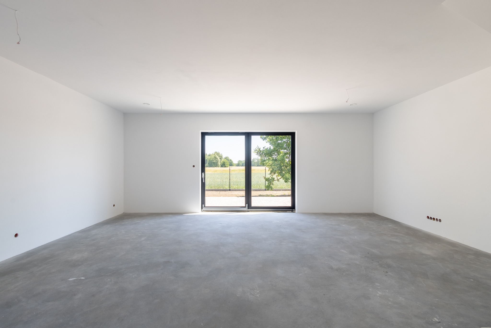 Surowe wnętrze z betonową podłogą i dużym oknem przesuwnym. Widoczne niedokończone instalacje elektryczne. Świeżo wylana podłoga.