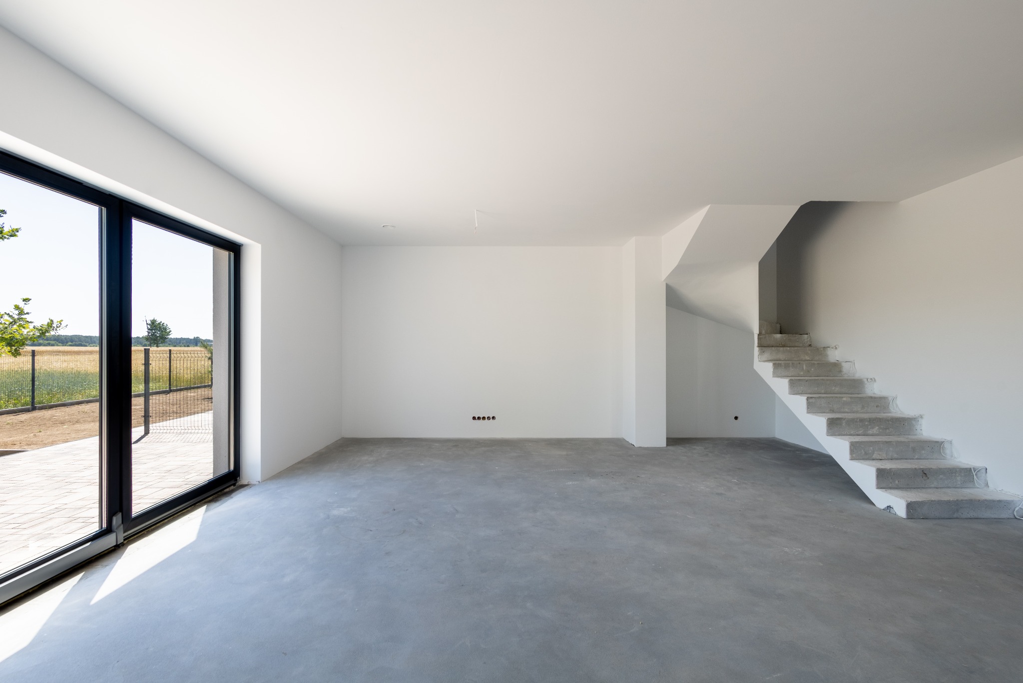 Surowe wnętrze: betonowa podłoga, białe ściany, schody w stanie surowym i duże okno z widokiem na zewnątrz. Widoczne gniazdka elektryczne. Wnętrze w trakcie wykańczania.