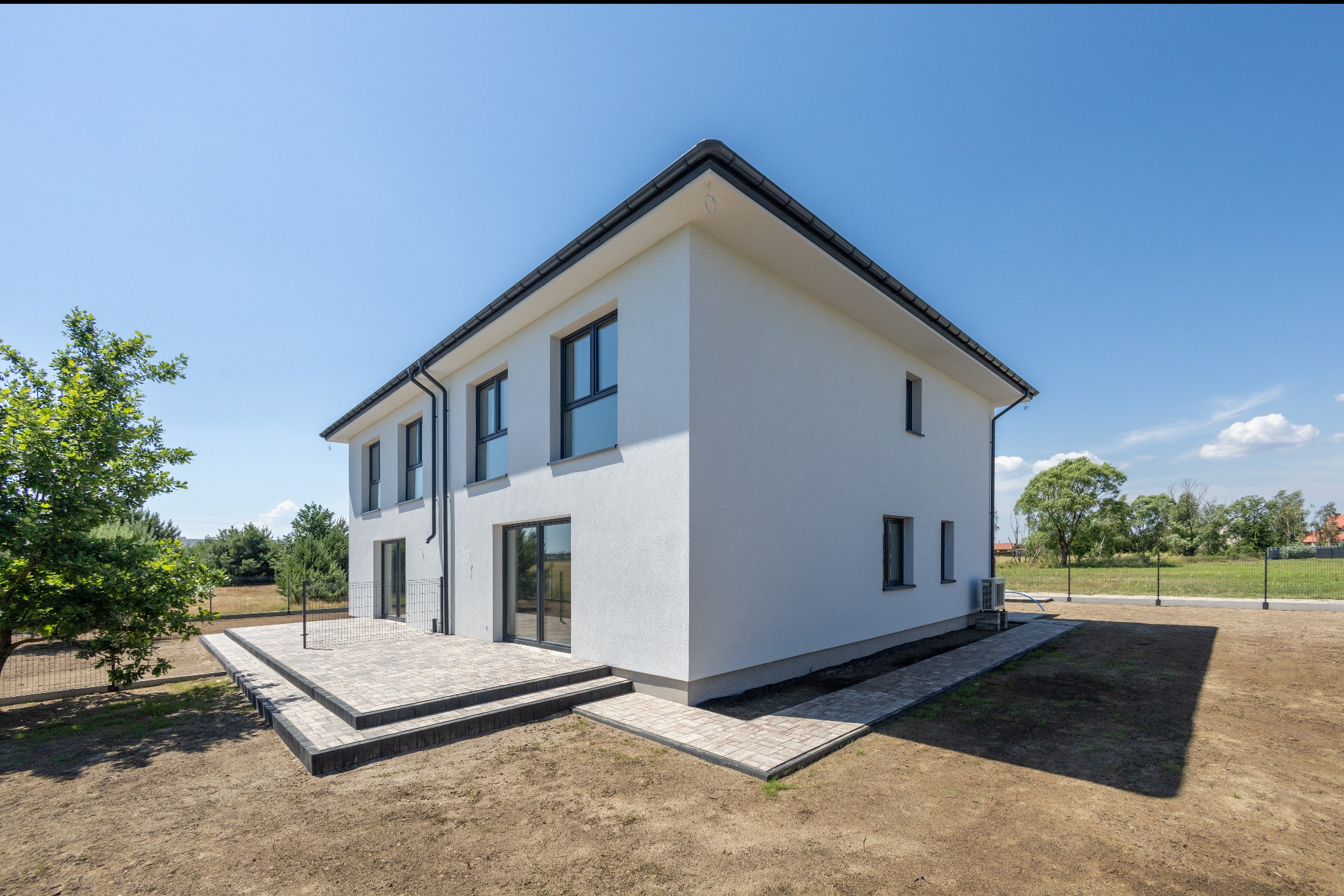 Nowoczesny, dwukondygnacyjny dom z białym tynkiem i ciemną stolarką okienną, otoczony trawnikiem i drzewami. Widoczny taras z kostki brukowej.