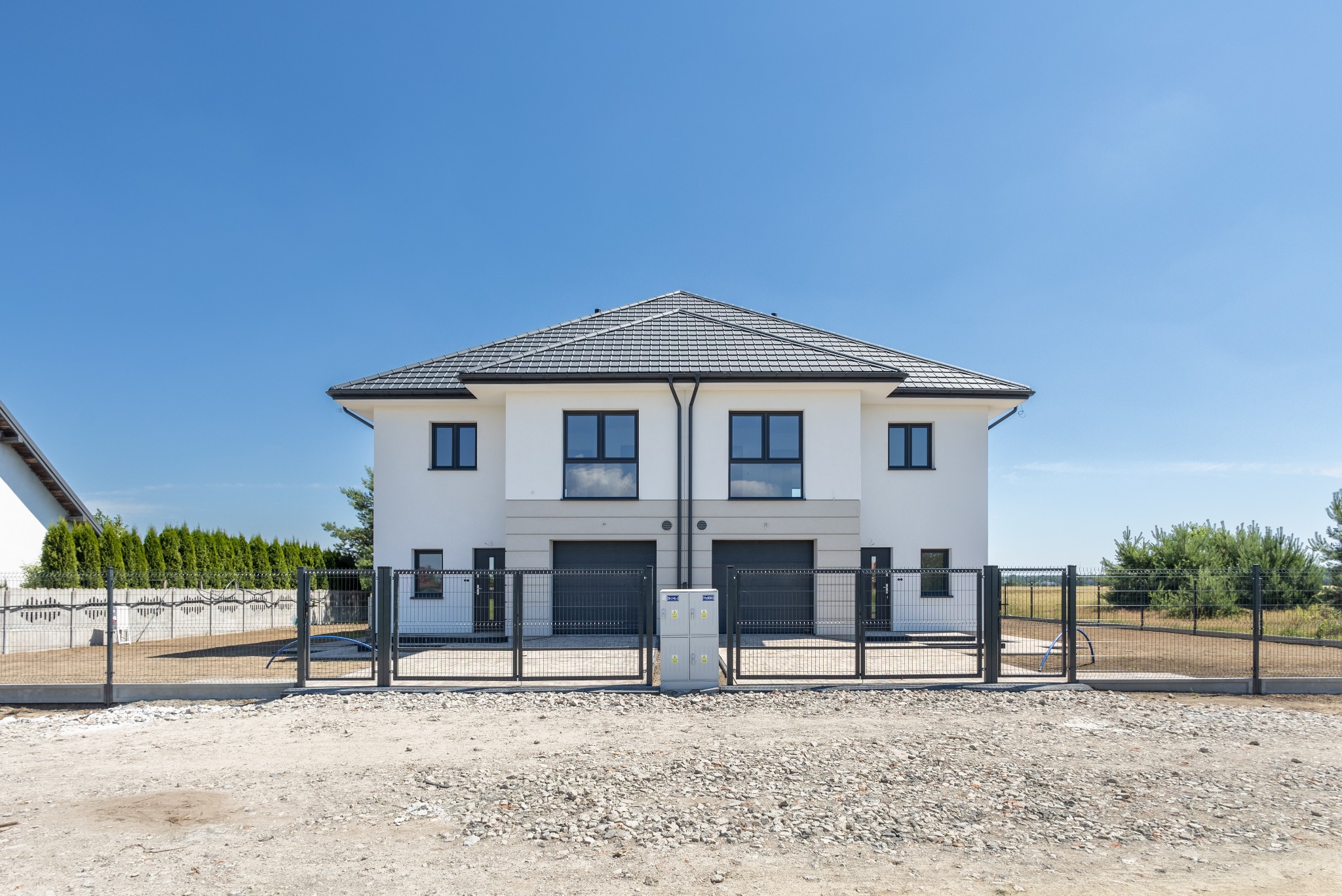 Nowoczesny, dwukondygnacyjny bliźniak z szarą dachówką i czarnymi oknami, ogrodzony metalową siatką na tle błękitnego nieba. Widoczne przyłącze elektryczne.