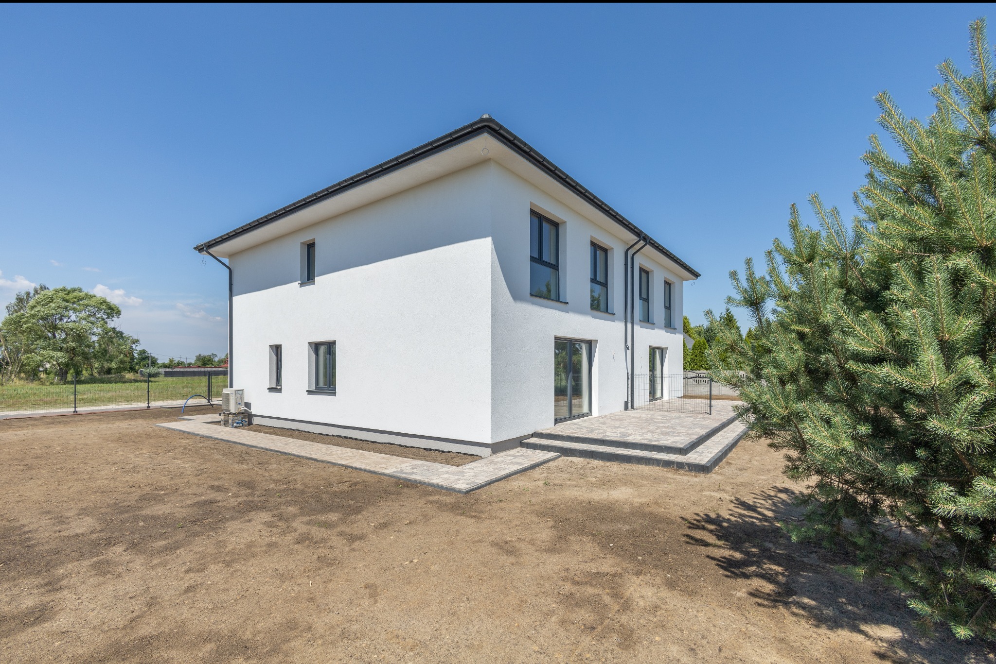 Nowoczesny, dwukondygnacyjny dom z białymi ścianami i ciemną stolarką okienną, otoczony surowym terenem i zielenią. Widoczny taras z kostki brukowej.