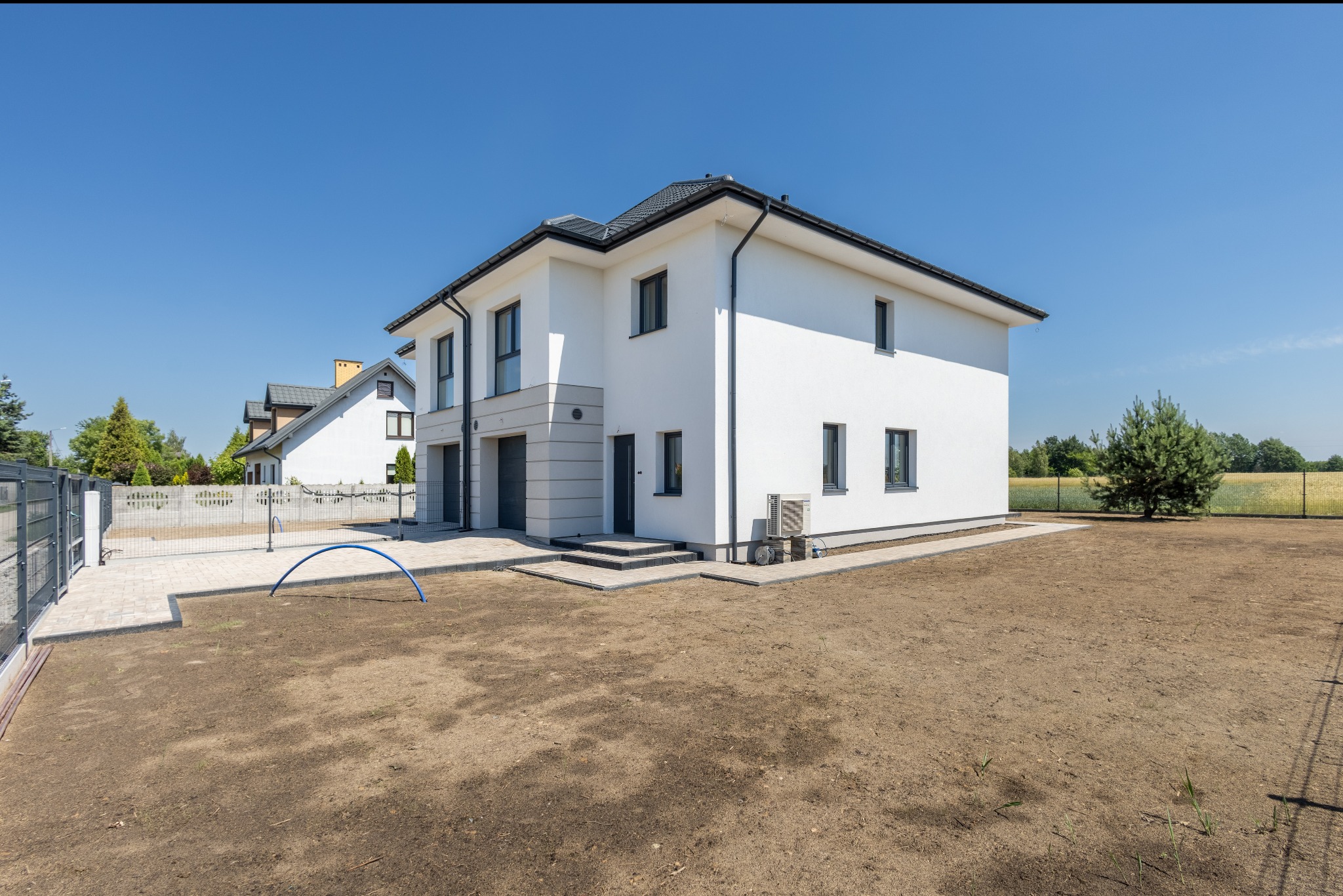 Nowoczesny, dwukondygnacyjny dom z białymi ścianami i szarymi oknami, widoczny z zewnątrz, z przygotowanym terenem pod ogród i błękitnym łukiem na trawniku.