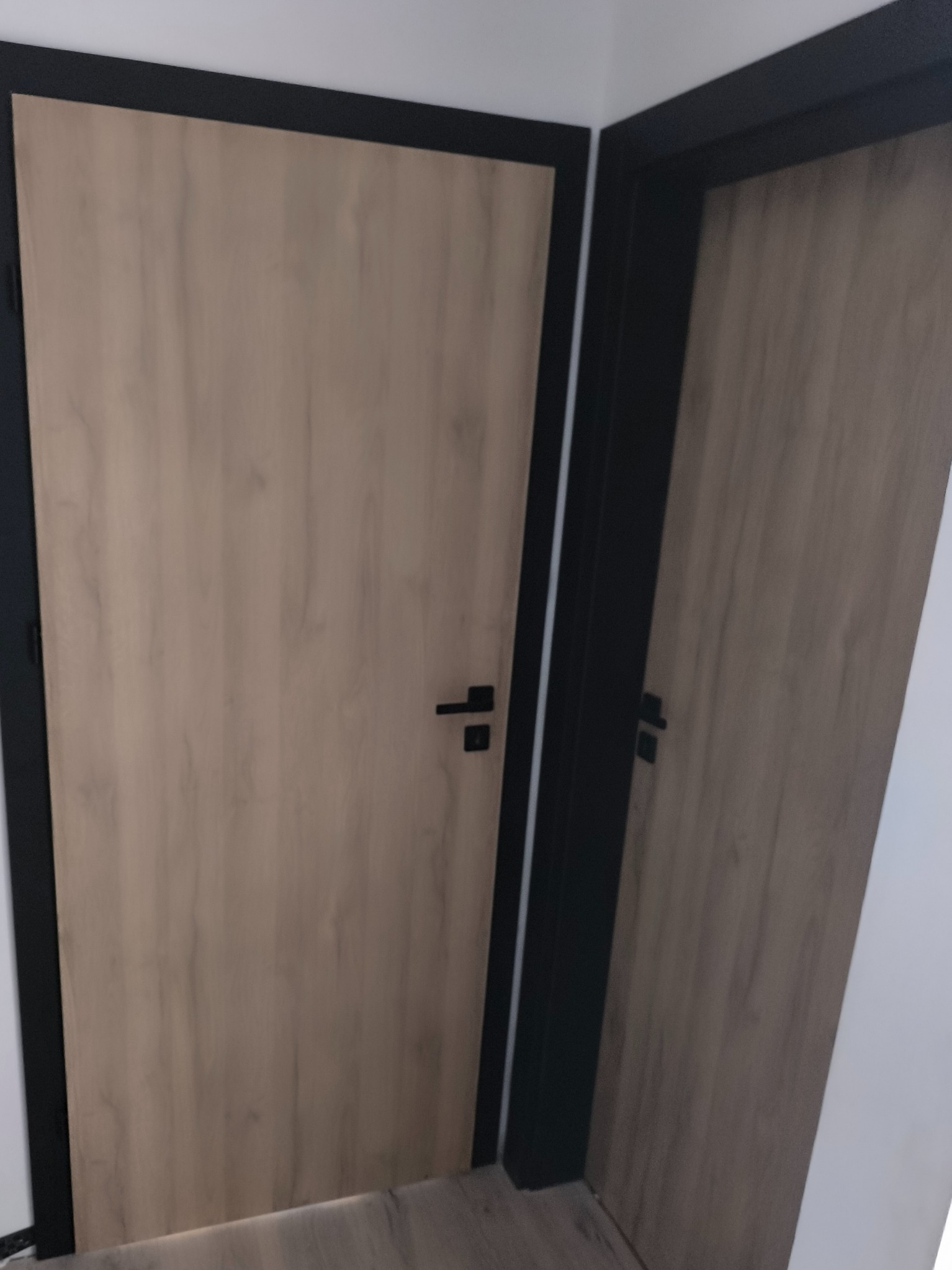 Dwie drewnopodobne drzwi wewnętrzne z czarnymi ościeżnicami i klamkami, widoczne z bliska, w rogu pomieszczenia. Minimalistyczny design.