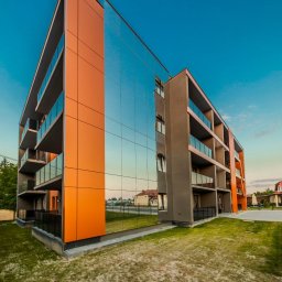 HulseyArt Projektowanie Wnętrz Katarzyna Mularska-Hulsey - Nowoczesny budynek apartamentowy z pomarańczowymi akcentami i szklaną fasadą odbijającą otoczenie, sfotografowany pod kątem, ukazujący detale wykończenia.