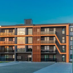 HulseyArt Projektowanie Wnętrz Katarzyna Mularska-Hulsey - Nowoczesny budynek apartamentowy z pomarańczowymi akcentami i dużymi oknami odbijającymi zachód słońca, widok z zewnątrz na tle błękitnego nieba.