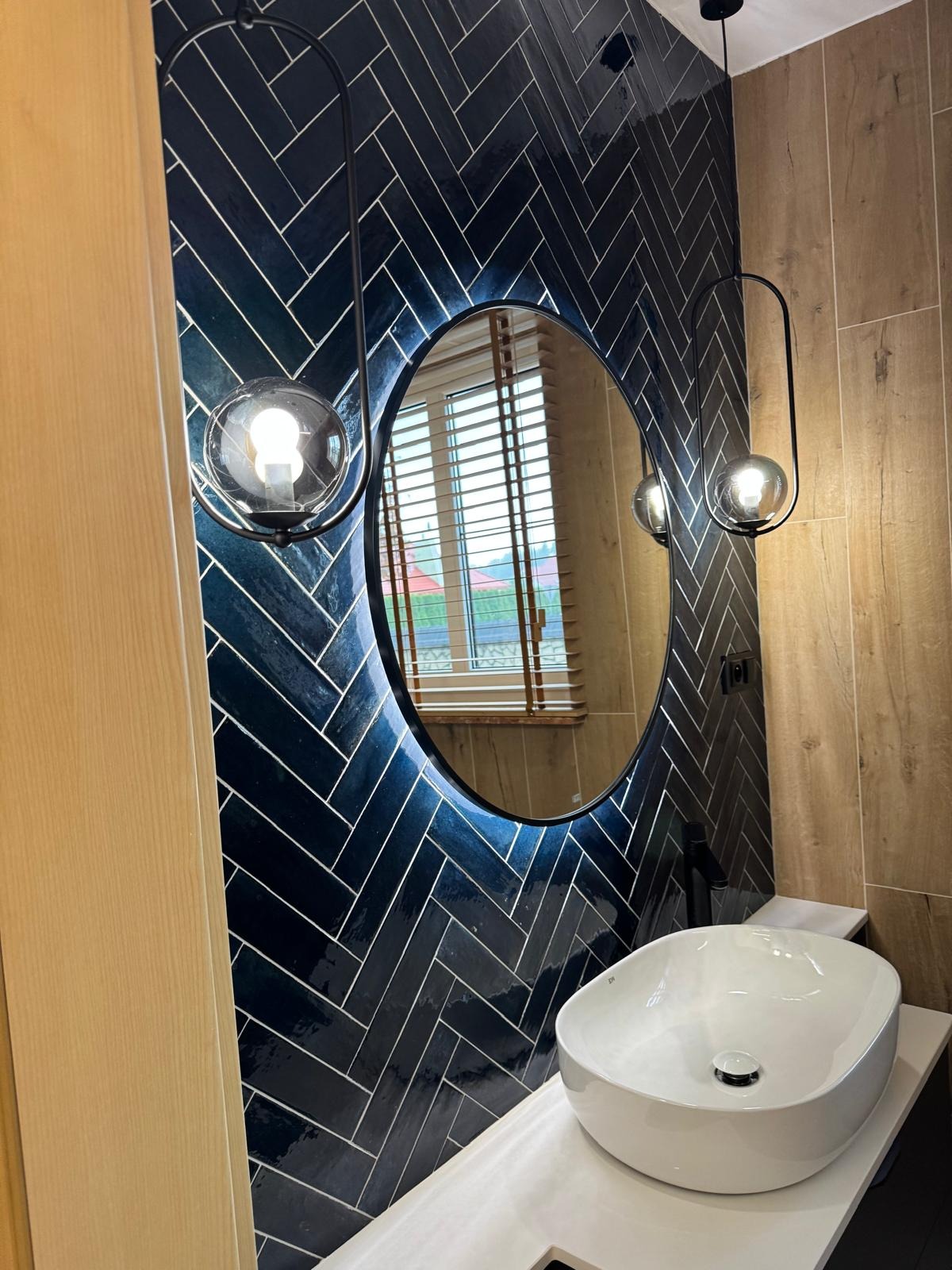 Nowoczesna łazienka z umywalką nablatową, owalnym lustrem z podświetleniem i ciemnymi płytkami ułożonymi w jodełkę. Widoczne lampy wiszące z kulistymi kloszami.