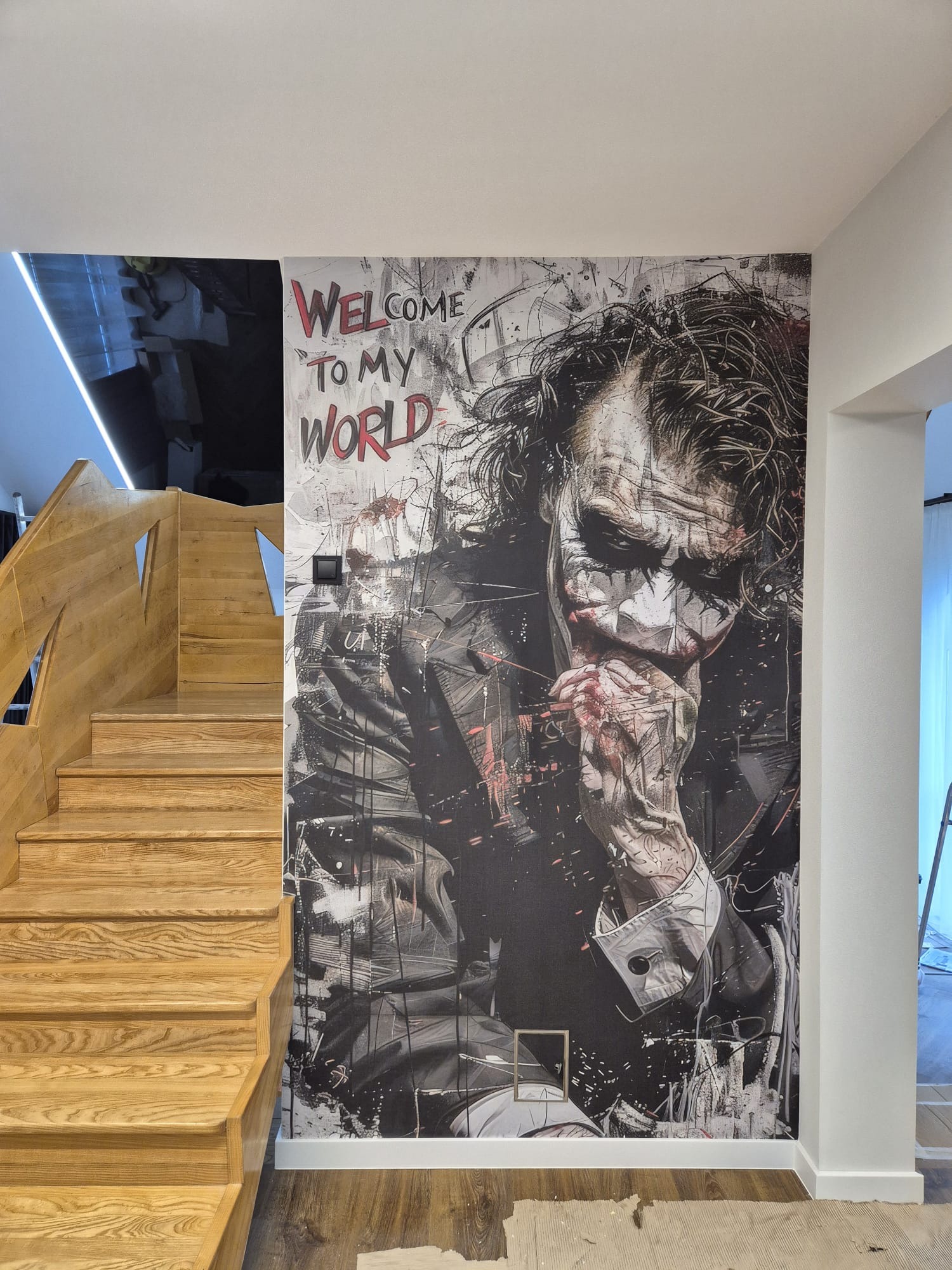 Klatka schodowa z drewnianymi schodami i fototapetą z wizerunkiem Jokera na ścianie. Nowoczesny design wnętrza z akcentem popkultury.