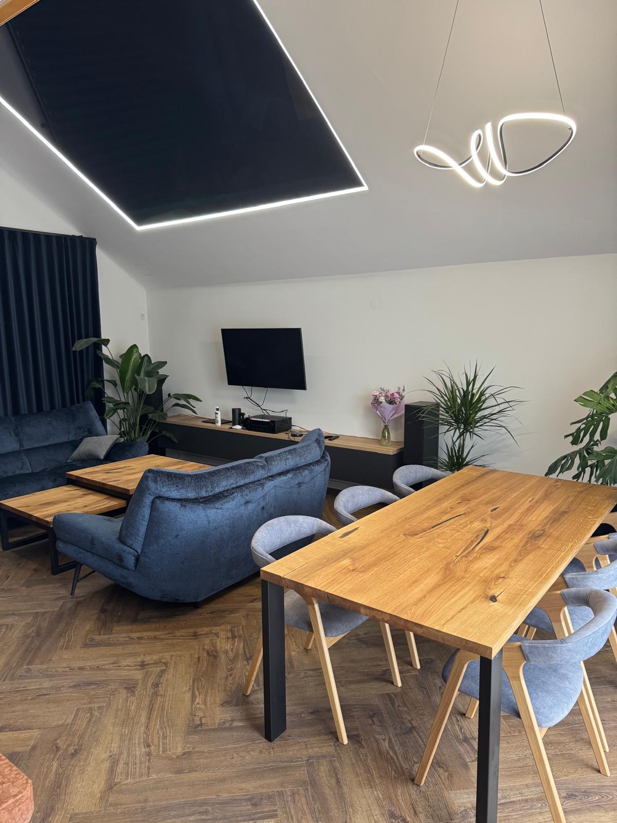 Nowoczesny salon z drewnianym stołem, niebieską sofą i parkietem w jodełkę. Czarny sufit z oświetleniem LED i designerska lampa wisząca. Rośliny doniczkowe dodają świeżości.