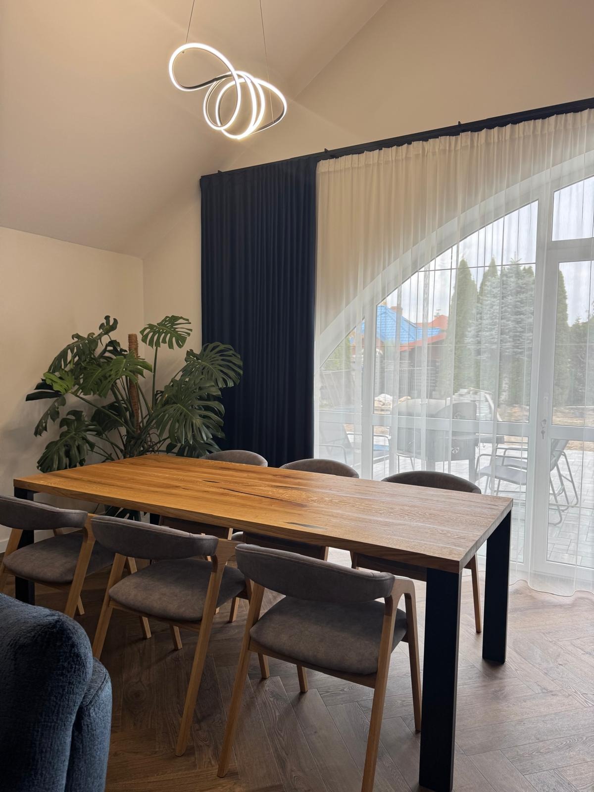 Stół z blatem z litego drewna i czarnymi nogami, otoczony szarymi krzesłami, w jasnym wnętrzu z rośliną Monstera i oknem z widokiem na ogród.