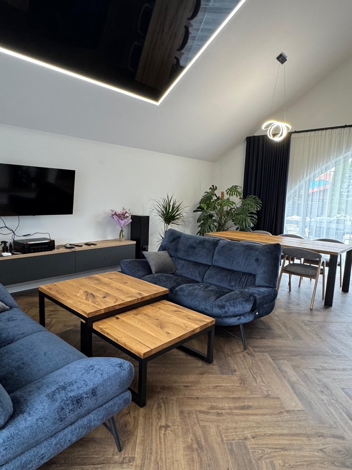 Salon z parkietem w jodełkę, niebieską sofą i drewnianymi stolikami kawowymi. Widoczne rośliny, stół jadalny i nowoczesne oświetlenie sufitowe.