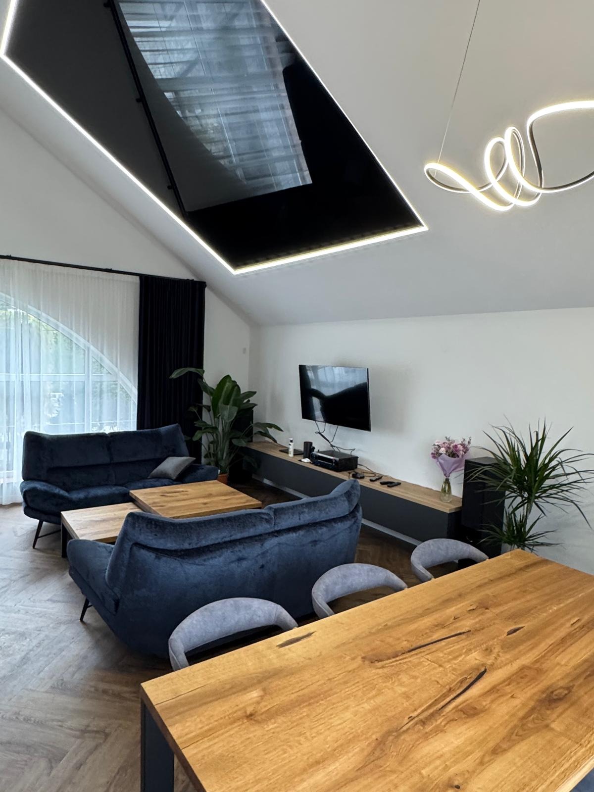 Salon z drewnianym stołem, sofą w odcieniach granatu i czarnym sufitem podwieszanym z oświetleniem LED. Nowoczesne wnętrze z roślinami doniczkowymi i telewizorem.