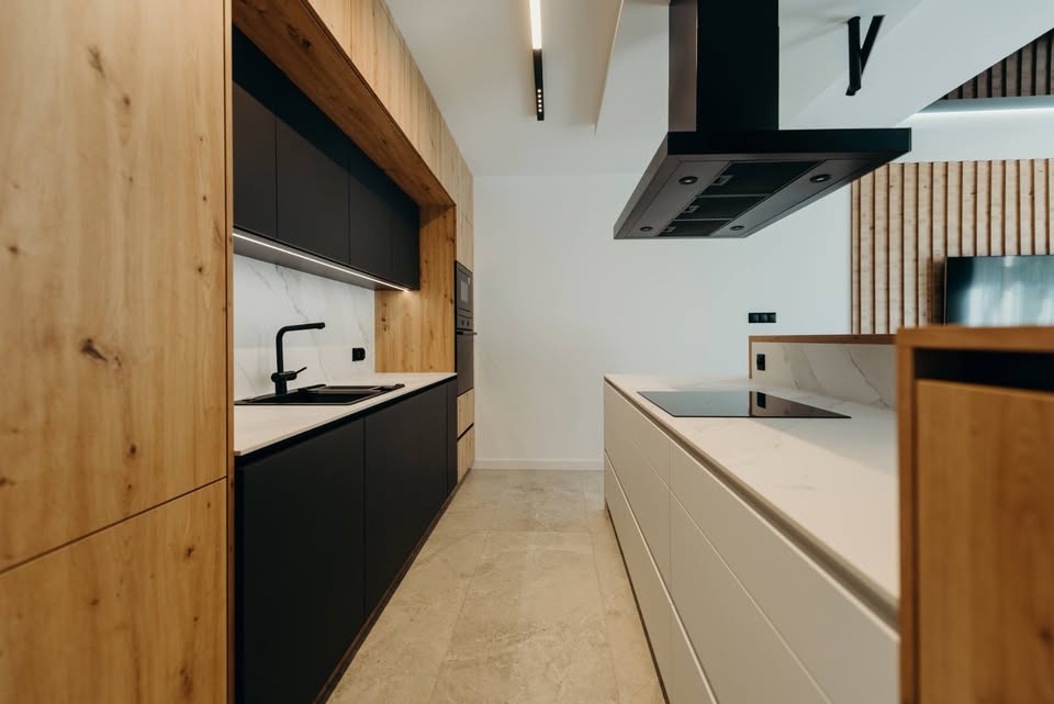 Nowoczesna kuchnia z drewnianymi i czarnymi szafkami, białym blatem i marmurową ścianą. Minimalistyczny design z oświetleniem LED i czarną armaturą.