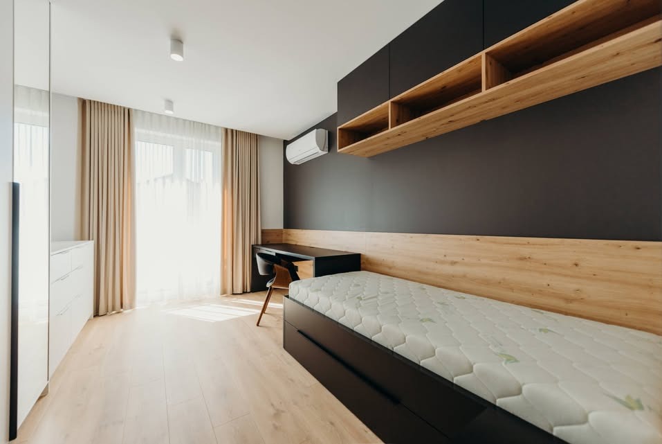 Nowoczesny pokój młodzieżowy z łóżkiem, biurkiem i półkami na ścianie. Jasne drewno i ciemny grafit tworzą spójną całość. Minimalistyczny design, naturalne światło.