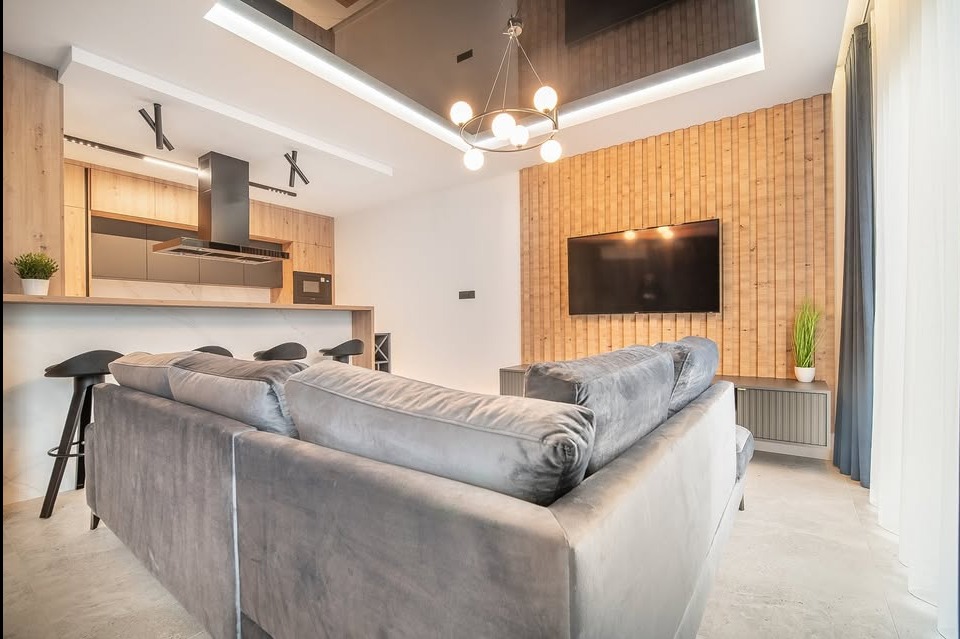 Nowoczesne wnętrze z drewnianą ścianą i szarą sofą. Widoczna kuchnia z wyspą i czarnymi hokerami. Całość oświetlona designerską lampą.