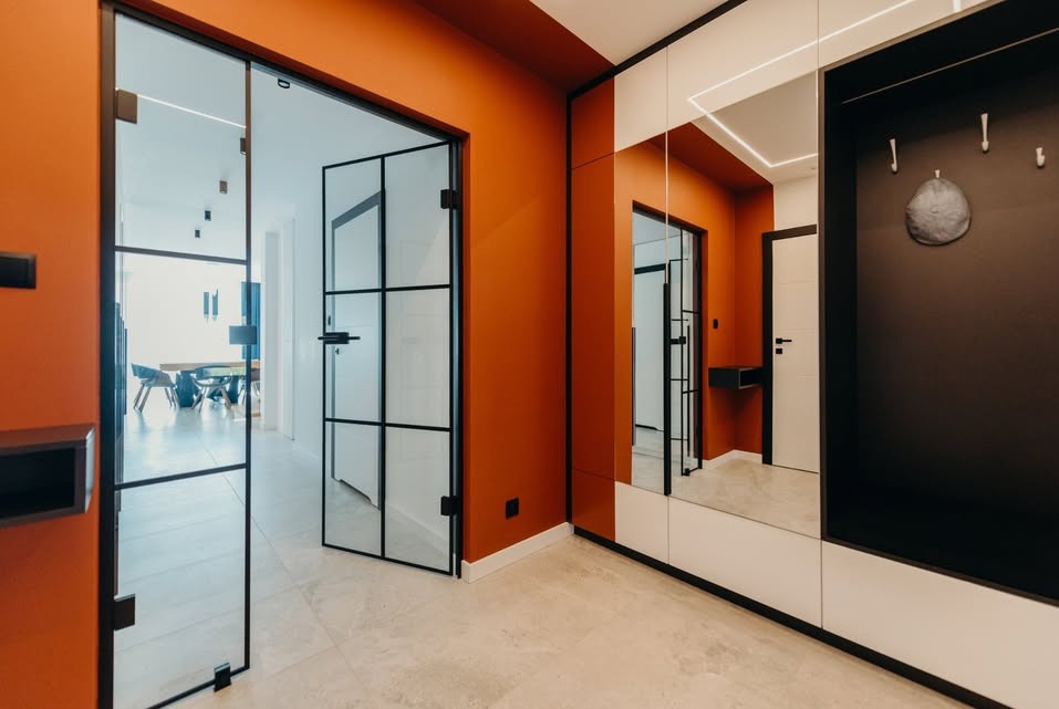 Nowoczesny przedpokój z pomarańczowymi ścianami, szklanymi drzwiami z czarnymi szprosami i szafą z lustrem. Widok na jadalnię w głębi.