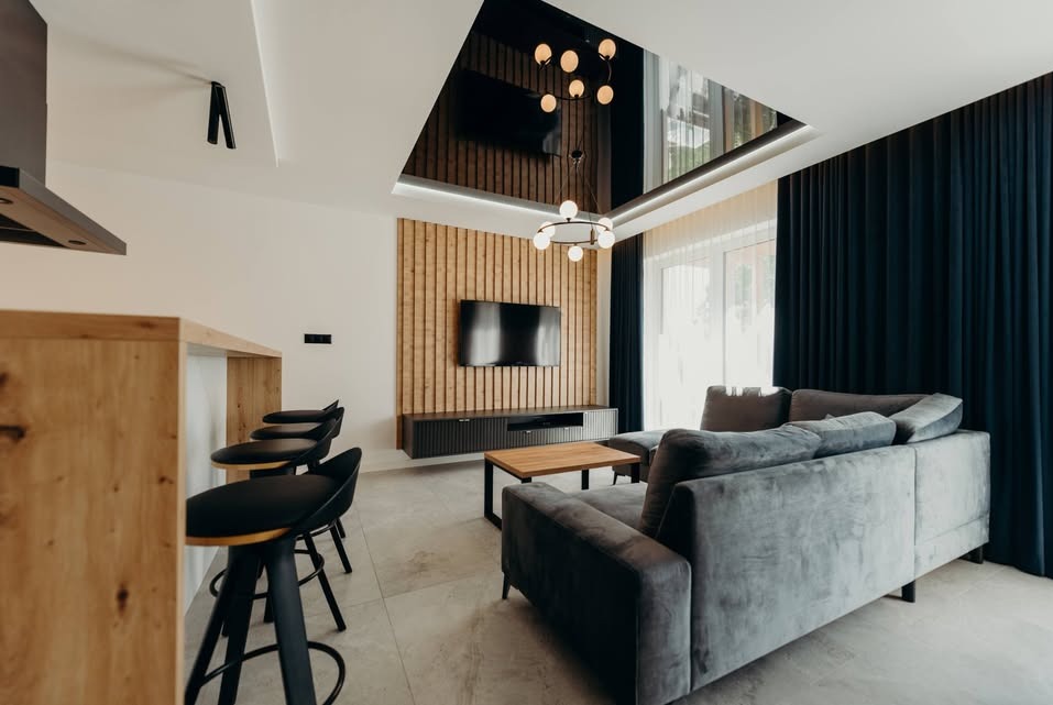 Nowoczesny salon z drewnianą ścianą TV, szarym narożnikiem i czarnymi hokerami przy drewnianym blacie. Lustrzane wykończenie sufitu z designerską lampą.