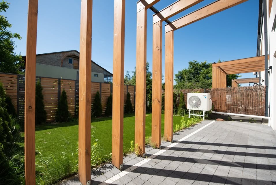 Nowoczesna pergola z drewna na tle zadbanego trawnika i domu. Widoczny klimatyzator zewnętrzny i drewniany płot. Cień rzucany przez konstrukcję tworzy geometryczny wzór na kostce.