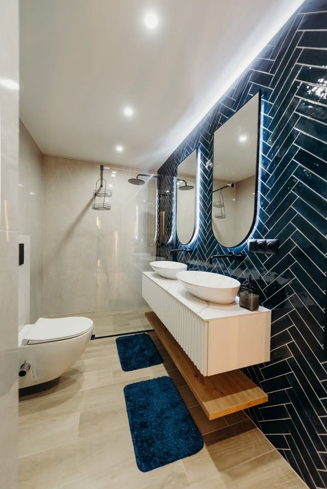 Nowoczesna łazienka z umywalkami nablatowymi, podświetlanymi lustrami i ścianą w ciemnej, wzorzystej glazurze. Uwagę zwracają detale wykończenia i oświetlenie.