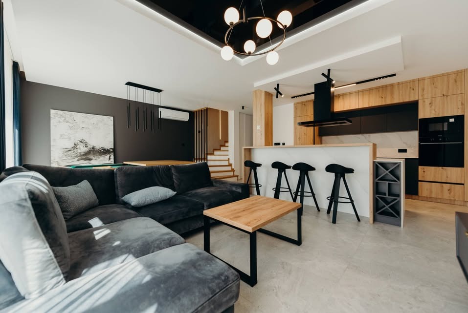 Nowoczesne wnętrze: szara sofa narożna, drewniany stolik kawowy, wyspa kuchenna z hokerami, drewniane szafki kuchenne i obraz na ścianie w odcieniach szarości.