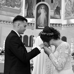 Strzyzewska_fotografia - Czarno-białe zdjęcie z ceremonii ślubnej w kościele: Panna młoda w koronie kwiatów całuje dłoń pana młodego w eleganckim garniturze na tle malowideł.
