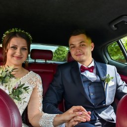 Strzyzewska_fotografia - Uśmiechnięta para młoda w samochodzie: panna młoda z bukietem i wiankiem, pan młody w garniturze z bordową muchą. Czerwona skórzana tapicerka.