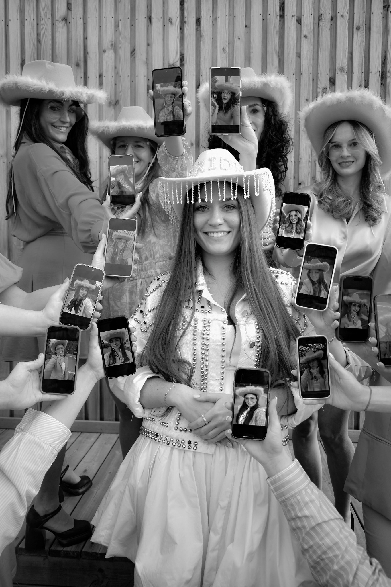 Czarno-białe zdjęcie grupy kobiet w kowbojskich kapeluszach, otaczających centralną postać, z telefonami prezentującymi zdjęcia portretowe w podobnej stylizacji.