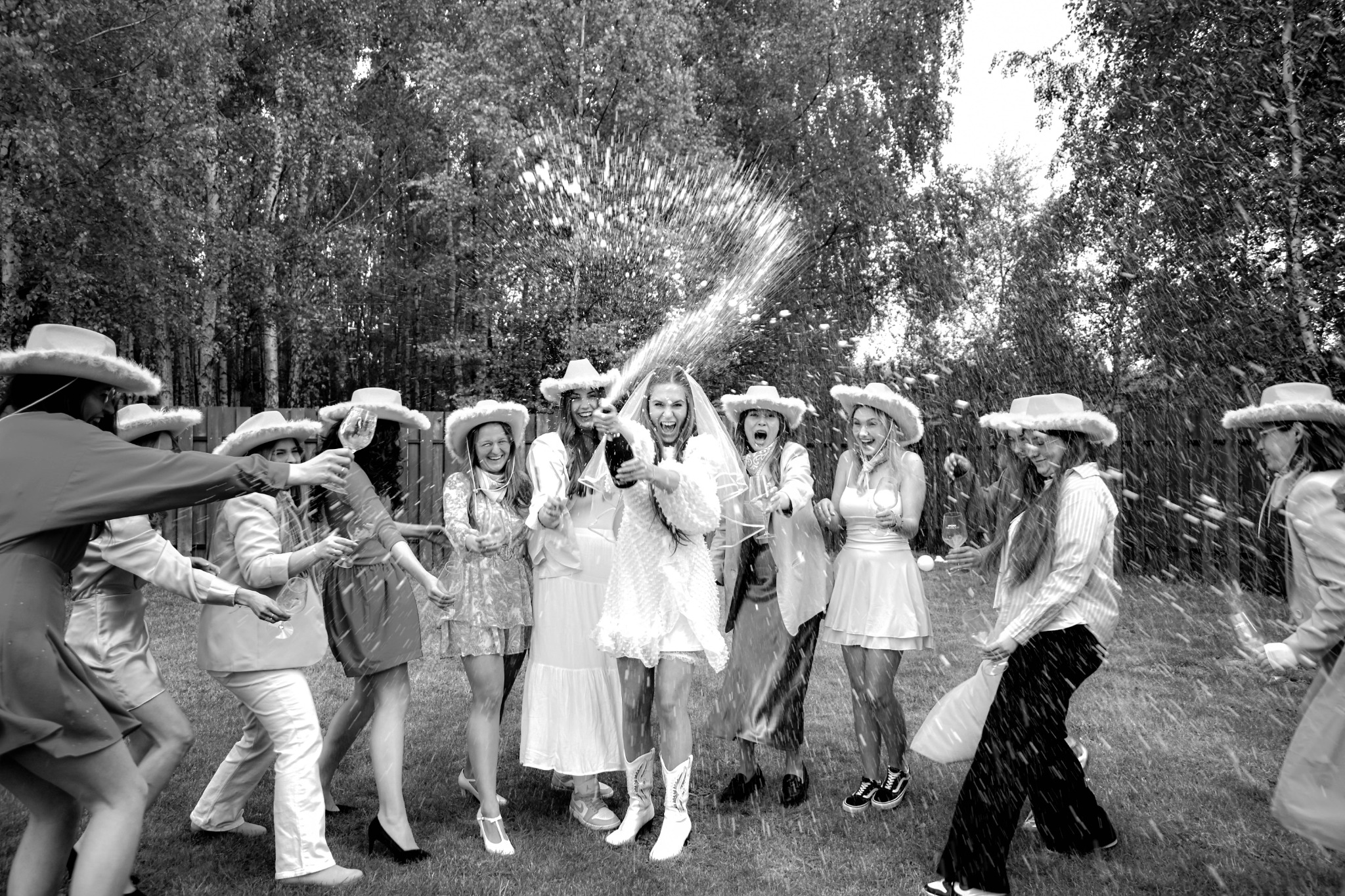 Czarno-białe zdjęcie panny młodej w kowbojskim kapeluszu i sukni, otoczonej przez druhny, otwierającej szampana na tle drzew podczas plenerowej sesji ślubnej.