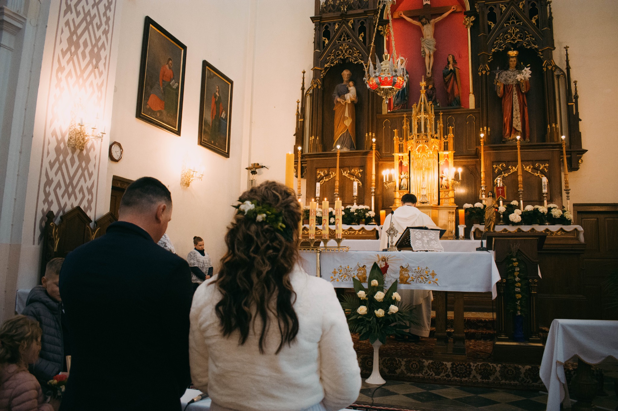 Para młoda podczas ceremonii ślubnej w kościele, widok z tyłu na pannę młodą z kwiatami we włosach, ołtarz z dekoracjami kwiatowymi i świecami w tle.