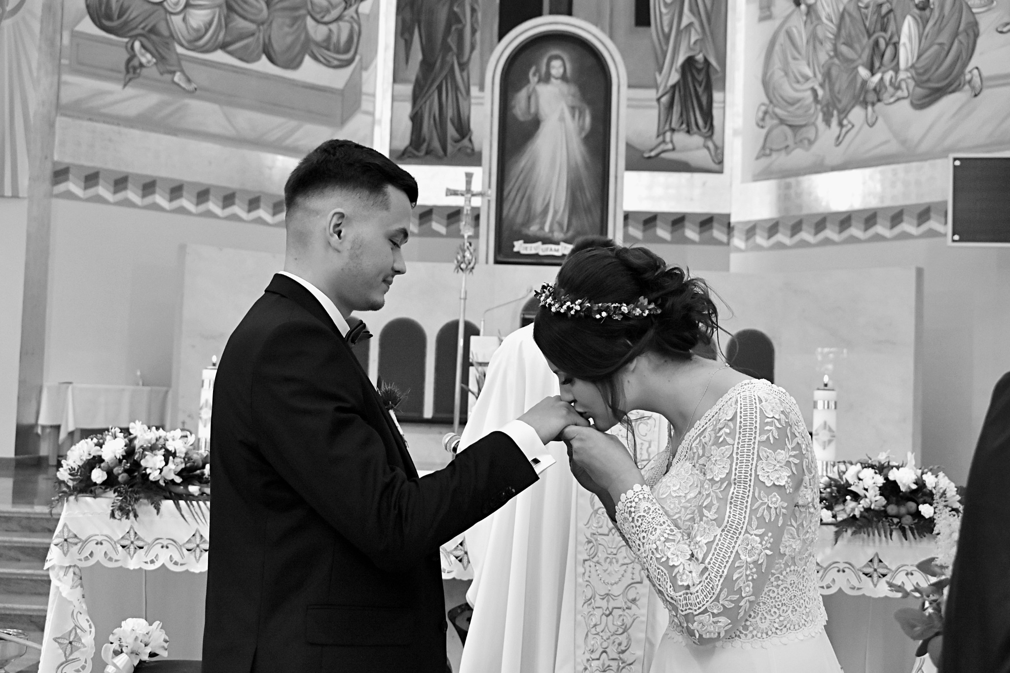 Czarno-białe zdjęcie z ceremonii ślubnej w kościele: Panna młoda w koronie kwiatów całuje dłoń pana młodego w eleganckim garniturze na tle malowideł.