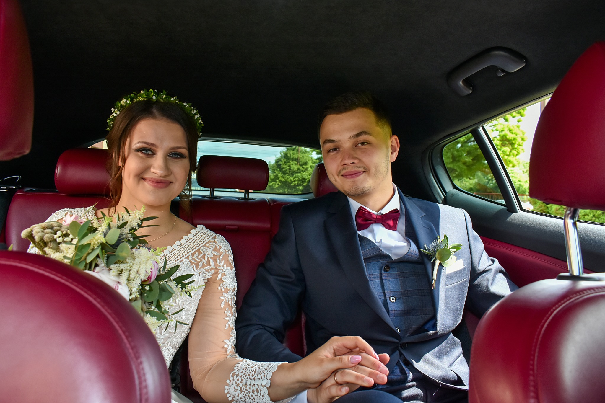 Uśmiechnięta para młoda w samochodzie: panna młoda z bukietem i wiankiem, pan młody w garniturze z bordową muchą. Czerwona skórzana tapicerka.