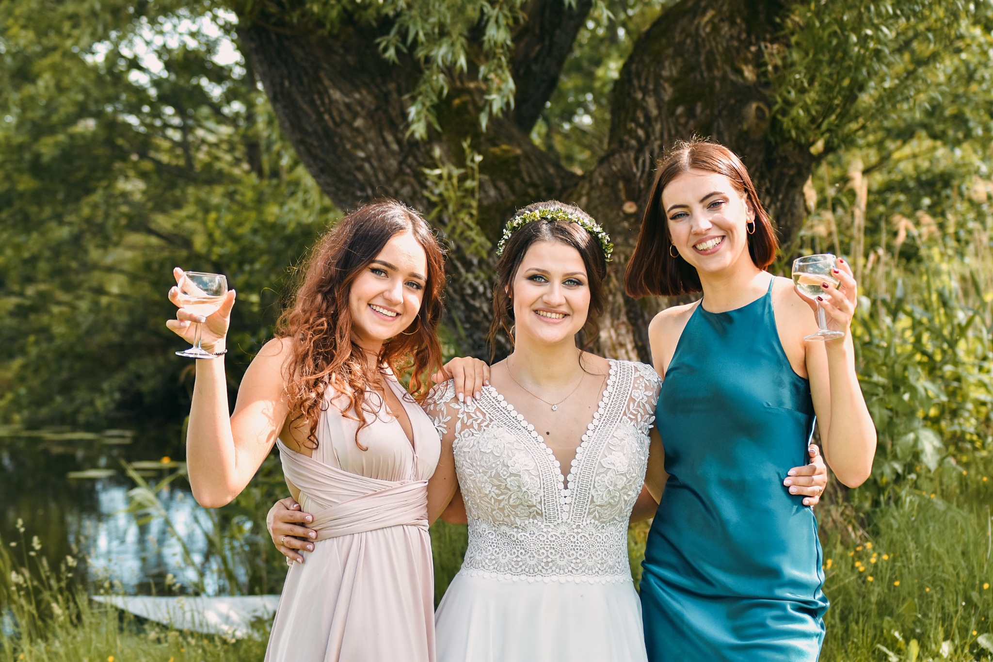 Uśmiechnięta panna młoda w koronie z kwiatów, otoczona dwiema druhnami w sukienkach, wznoszą toast na tle zieleni i wody. Słoneczny dzień.