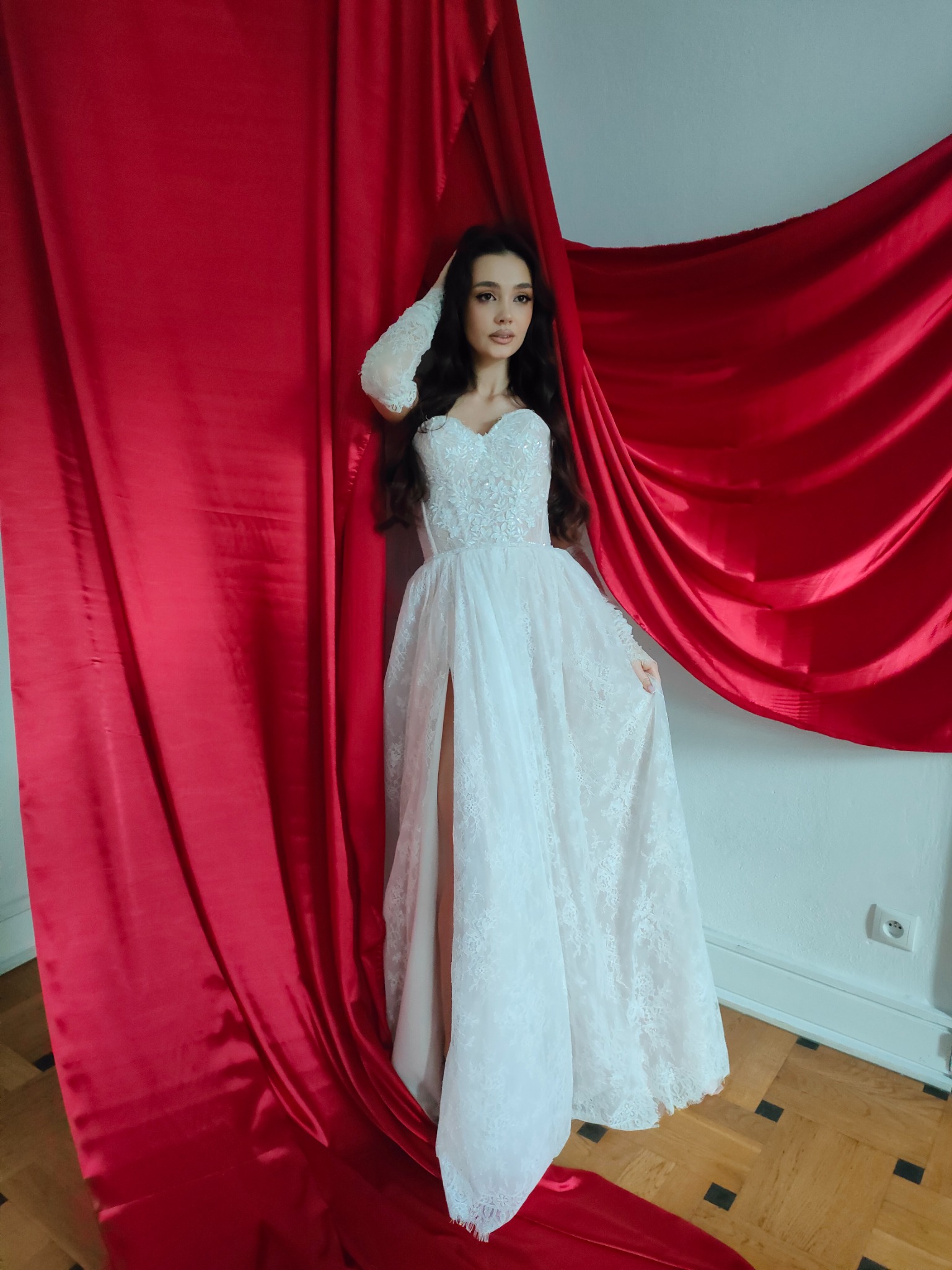 Kobieta w koronkowej sukni ślubnej z rozcięciem, pozująca na tle czerwonej, błyszczącej tkaniny. Ujęcie w studio, z widocznym fragmentem podłogi i gniazdka.